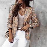 Frau in kariertem Blazer, weißer Hose und Hut lehnt an Betonwand. Modischer Look mit Schmuck und Handtasche. Eleganter Streetstyle für Herbst.