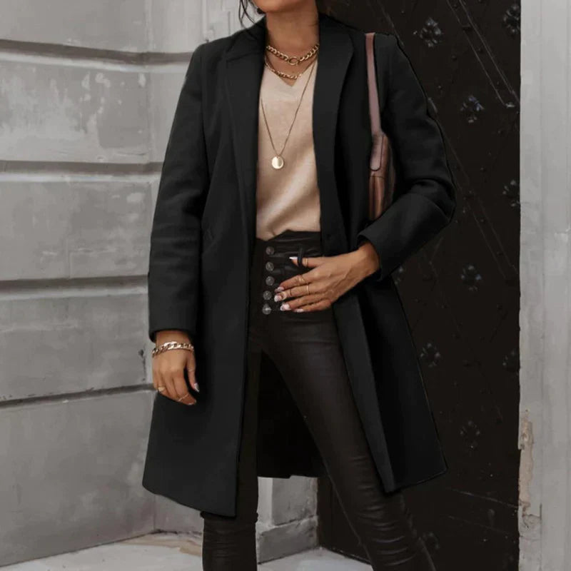 Frau in schwarzem Mantel und Lederhose vor dunkler Tür, trägt goldene Halskette und Armbänder, modisches Herbst-Outfit, elegante Damenmode.