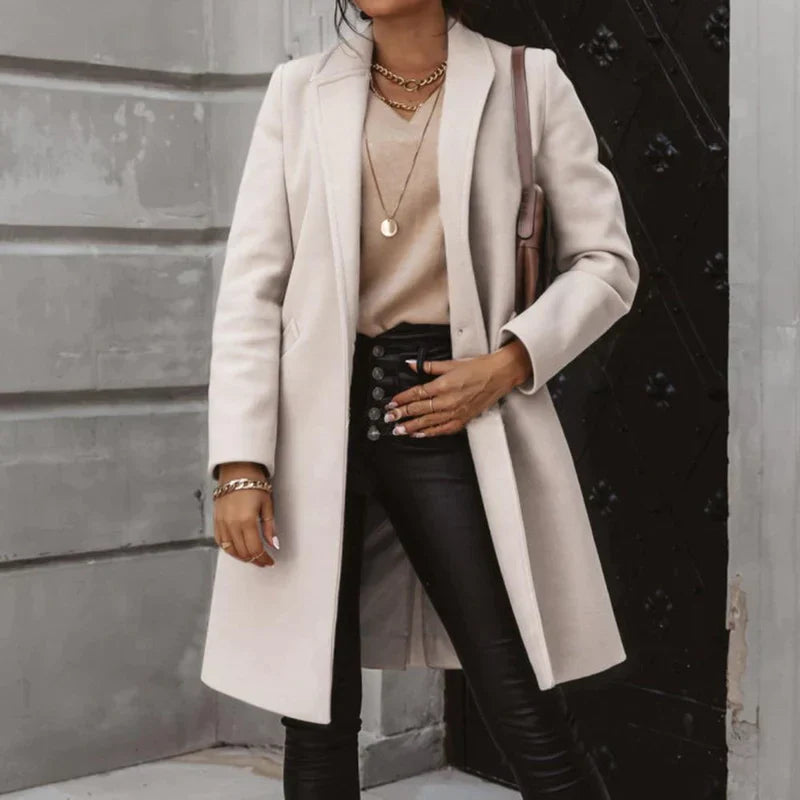 Dame in elegantem beigem Mantel, schwarzer Lederhose und goldenen Accessoires vor einer grauen Wand. Modetrend Herbst, stilvolle Damenmode.