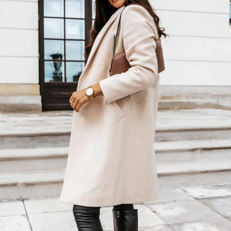 Dame in elegantem, beigem Wollmantel, schwarze Lederhose und Stiefel, braune Handtasche, vor einem Gebäude mit weißen Wänden und dunkler Tür.