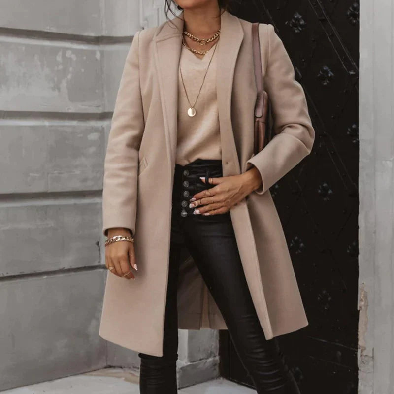 Frau in elegantem beigem Mantel, kombiniert mit schwarzer Lederhose und goldenen Accessoires, steht vor einer grauen Wand. Modischer Herbst-Look.