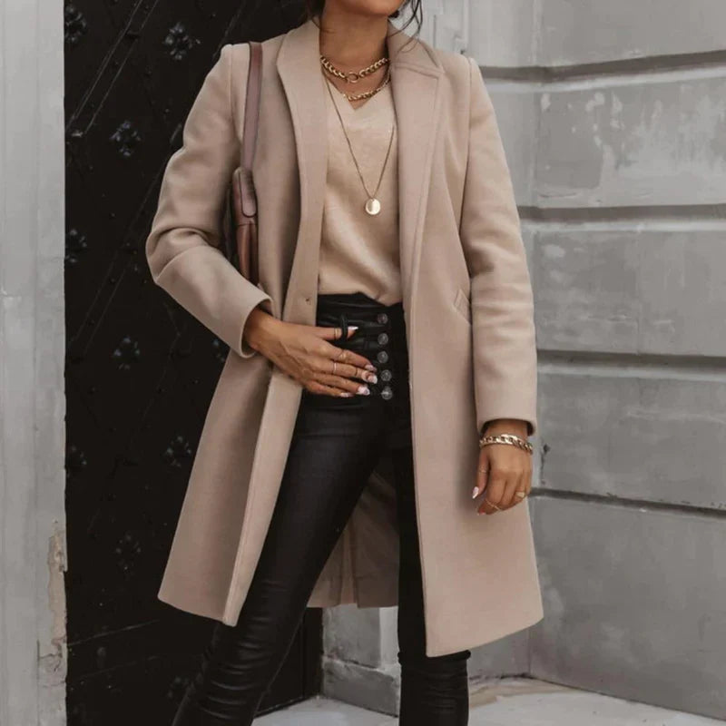 Frau in beigem Mantel, schwarzer Lederhose und goldenen Accessoires vor grauer Wand. Elegante Herbstmode, stilvoller Look, Streetstyle.
