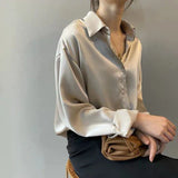 Dame in eleganter, beiger Seidenbluse, sitzt mit verschränkten Armen. Modetrend, stilvolle Damenbekleidung, luxuriöse Stoffe, minimalistischer Look.