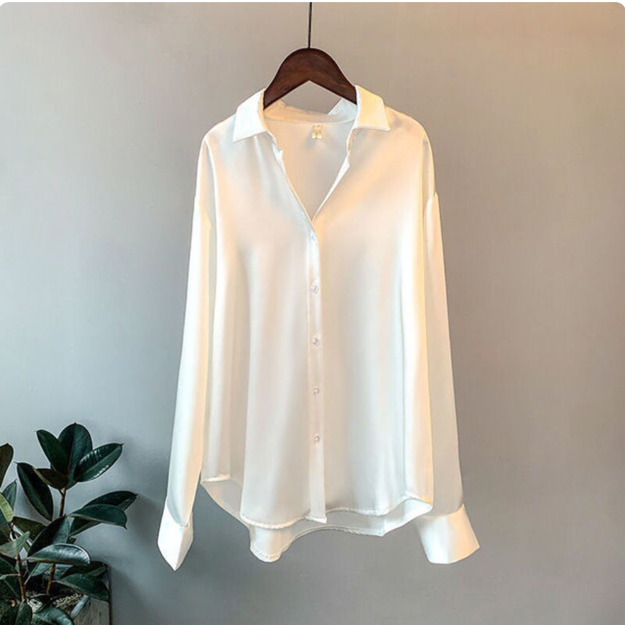 Weiße Damenbluse aus Seide auf Holzkleiderbügel, minimalistisch, elegant, langärmlig, vor grauem Hintergrund, neben grüner Zimmerpflanze.