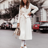 Eine Frau in einem eleganten weißen Mantel steht auf einer städtischen Straße, hält eine beige Handtasche. Stilvolle Herbstmode, urbane Kulisse.